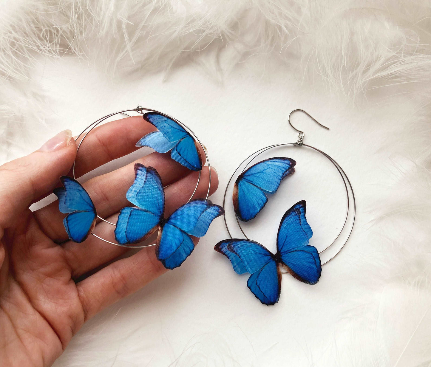 Hand Holds Silver Hoop Earrings with 3D Blue Butterflies on White Feathers Background
