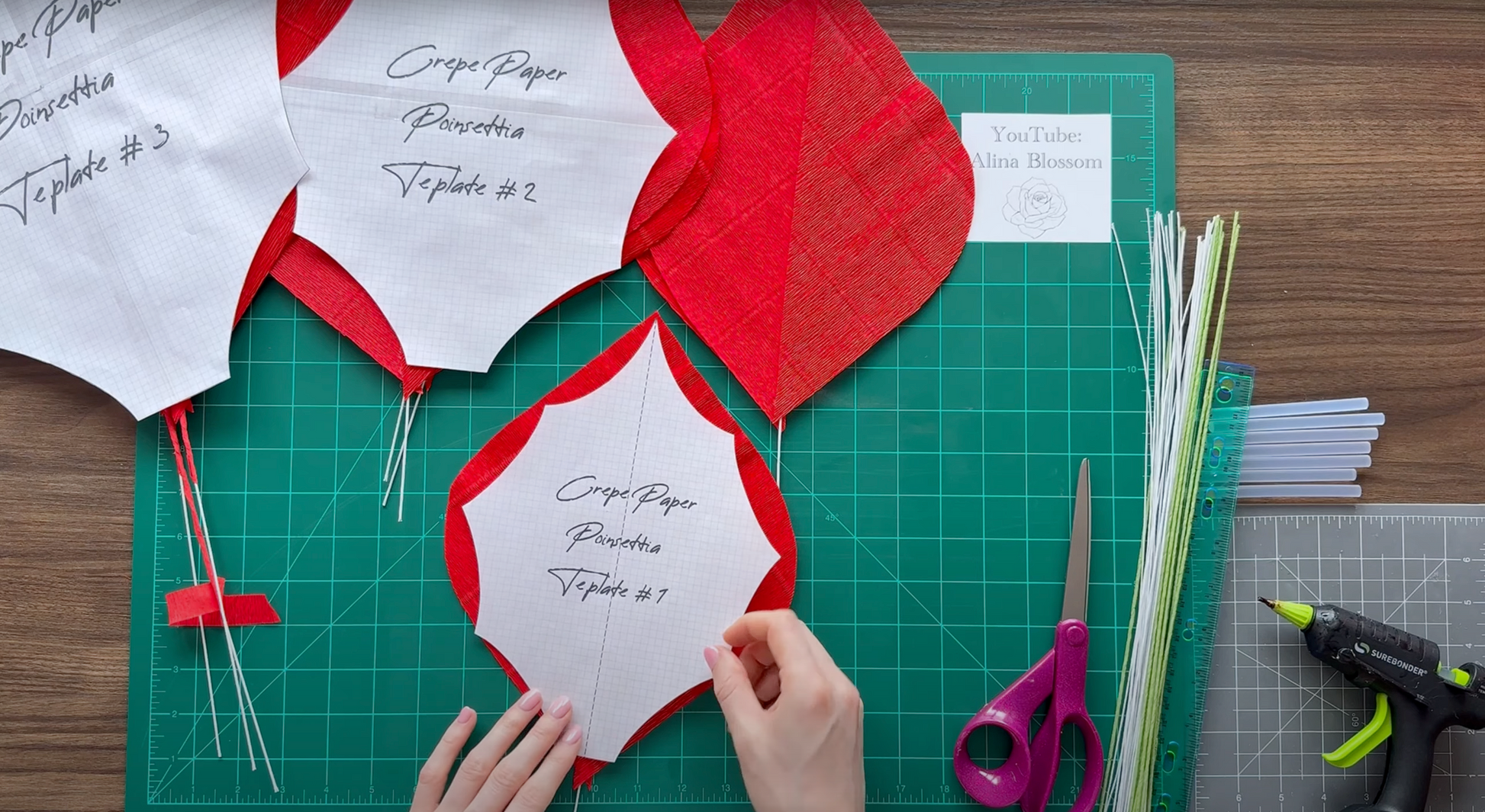 Hands working with red and white paper cutouts on a green cutting mat with scissors and a hot glue gun.