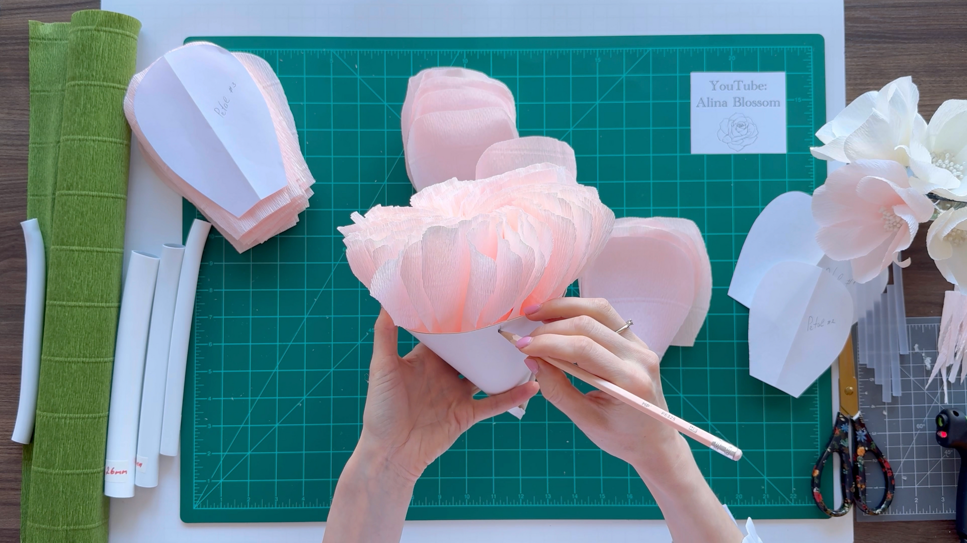 Alina Blossom holding a pink paper flower craft on a green cutting mat with scissors and other crafting materials.