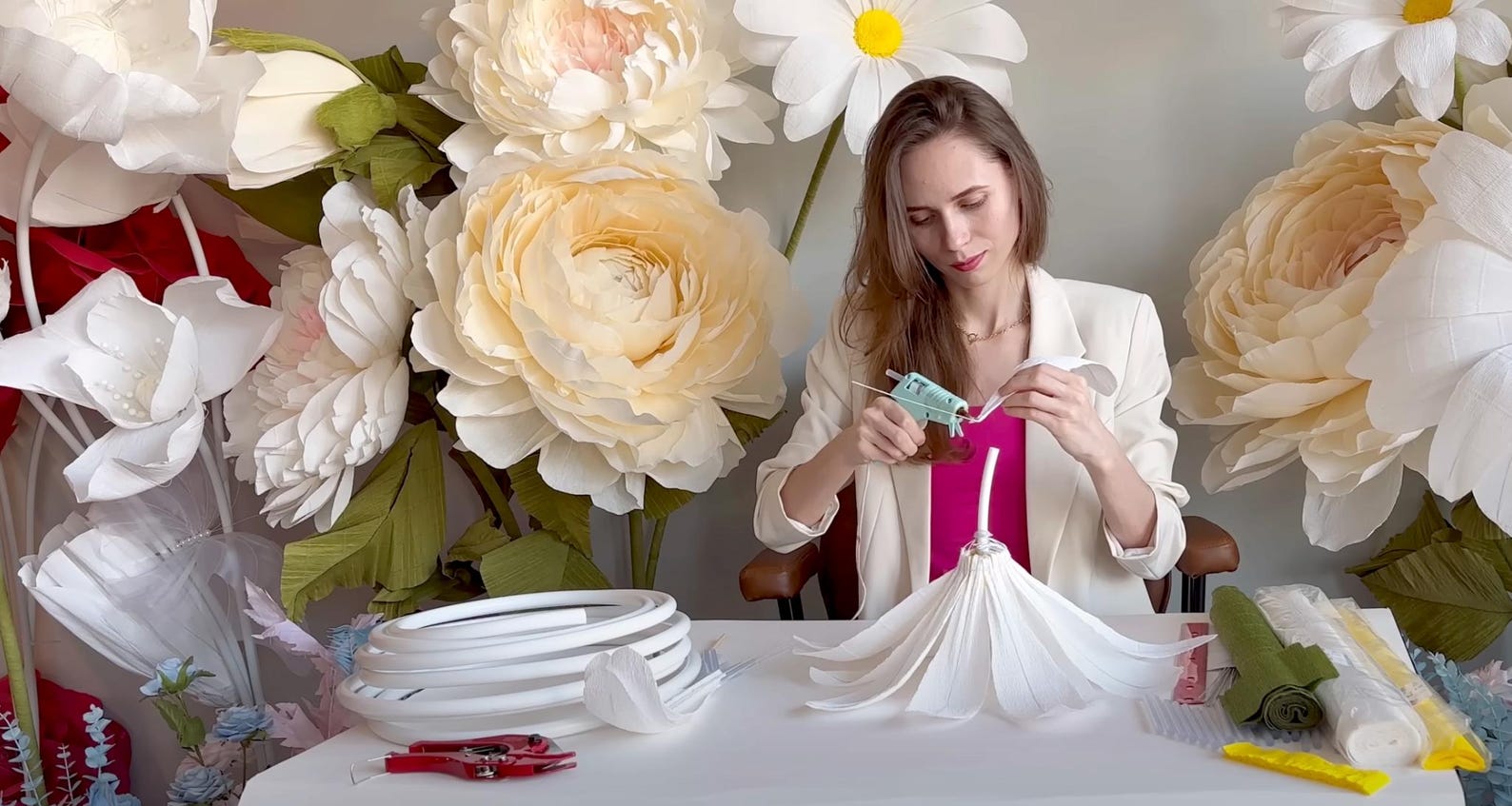 Alina Blossom arranging flowers in a decorative vase with large artificial flowers in the background