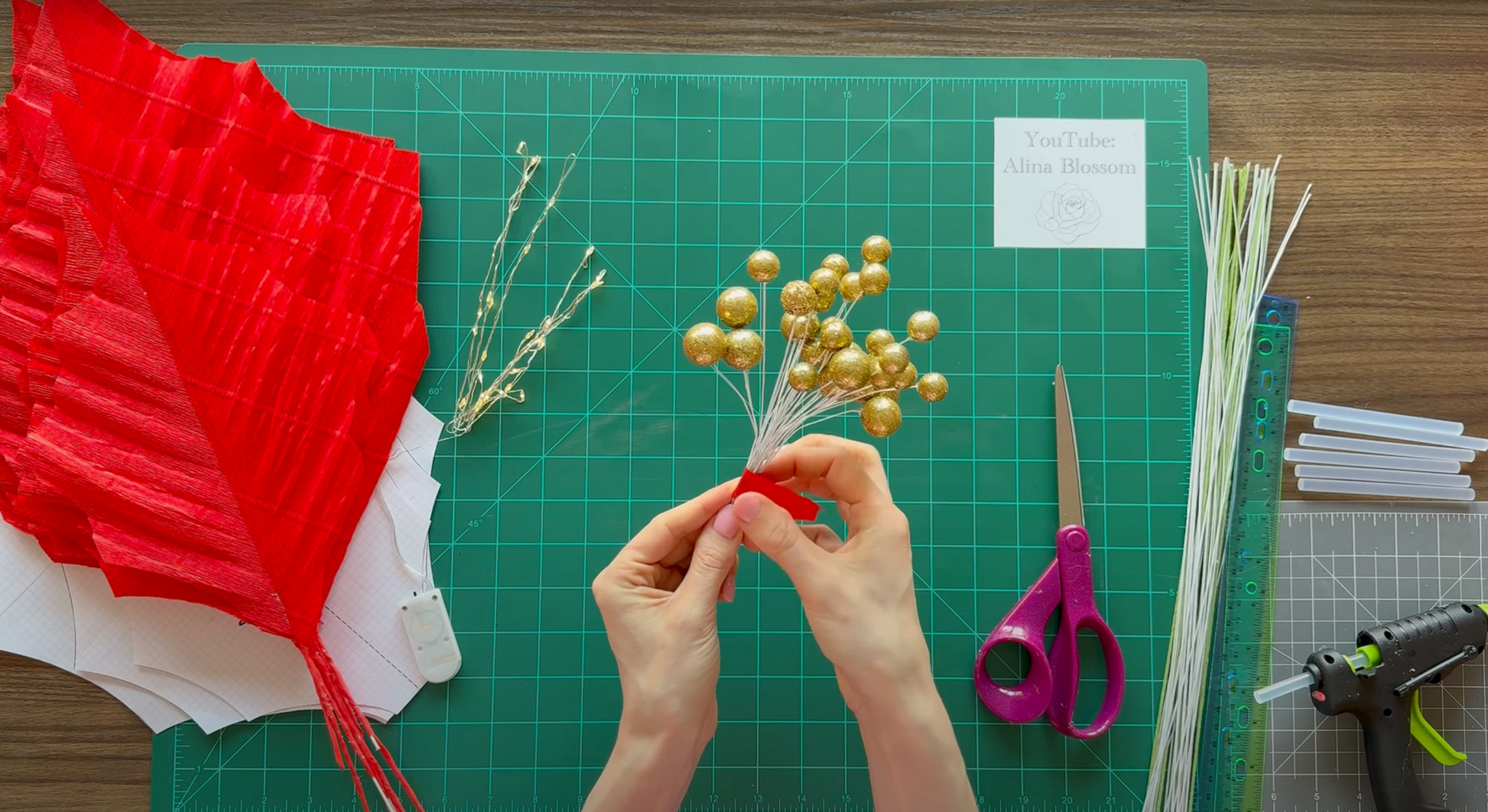 Hands working with red paper, gold decorations, and scissors on a green cutting mat.