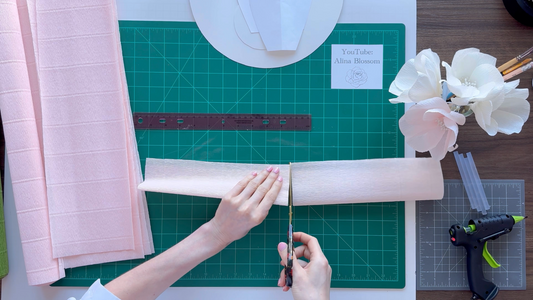 Alina Blossom cutting pink paper with scissors on a green cutting mat, surrounded by crafting tools.