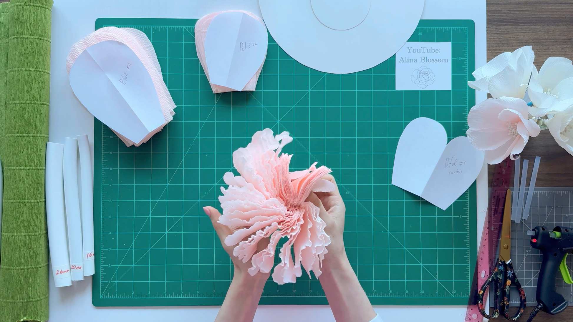 Alina Blossom crafting a pink paper flower on a green cutting mat with various craft materials.