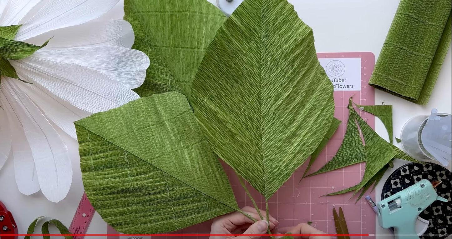 Green paper leaves on a pink cutting mat with white flowers and crafting tools in the background.