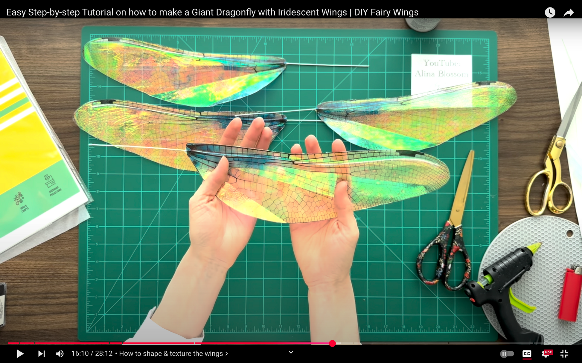 Hands holding iridescent fairy wings on a cutting mat with crafting tools.