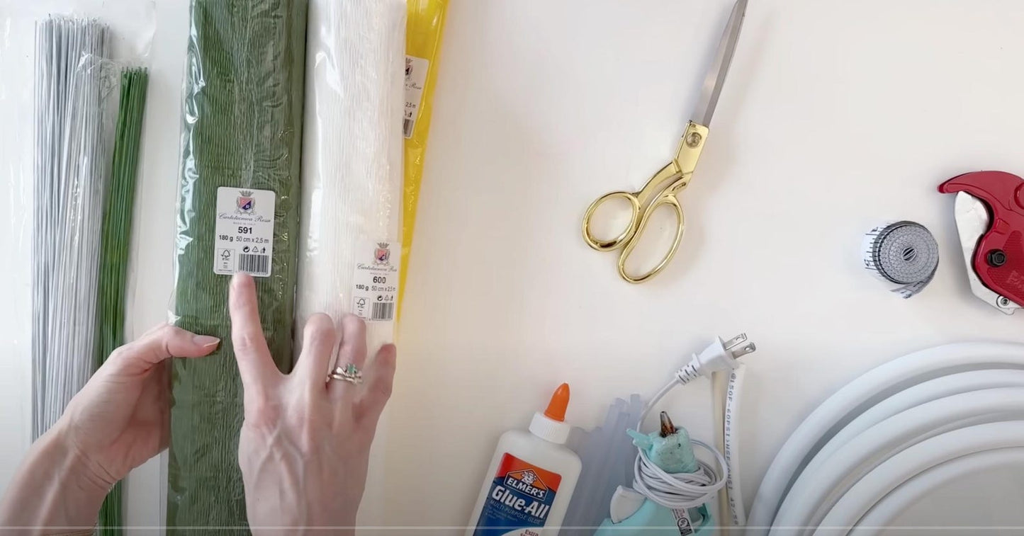 Hands holding a roll of green paper with craft supplies on a white surface.