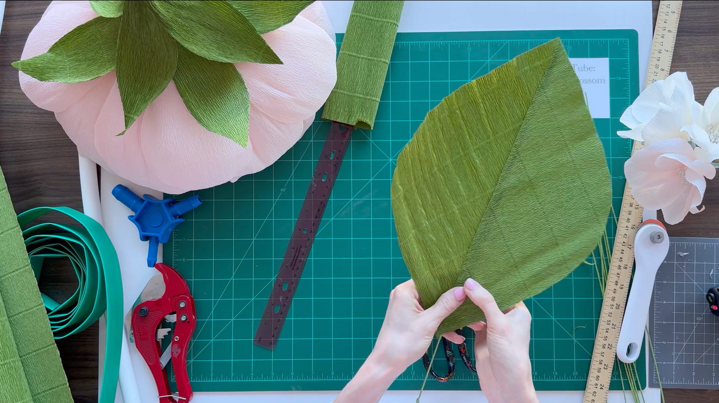 Hands working with green fabric leaves on a cutting mat with crafting tools.