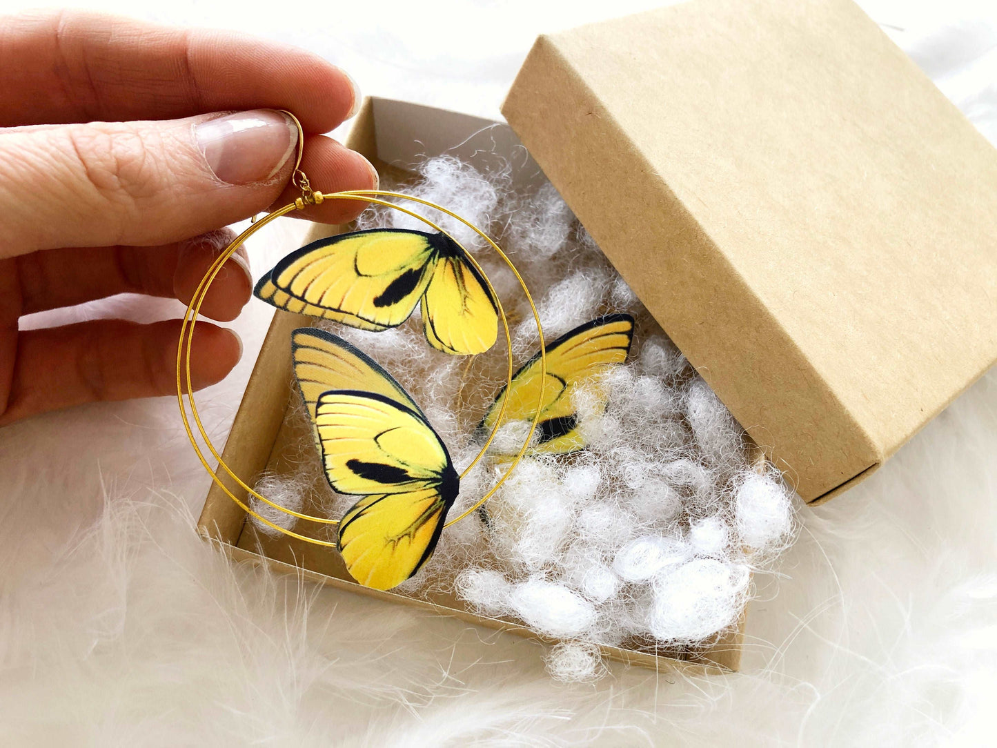 Hand Holds Double Hoop Earrings with Yellow Butterflies in Craft Box on White Background and Feathers