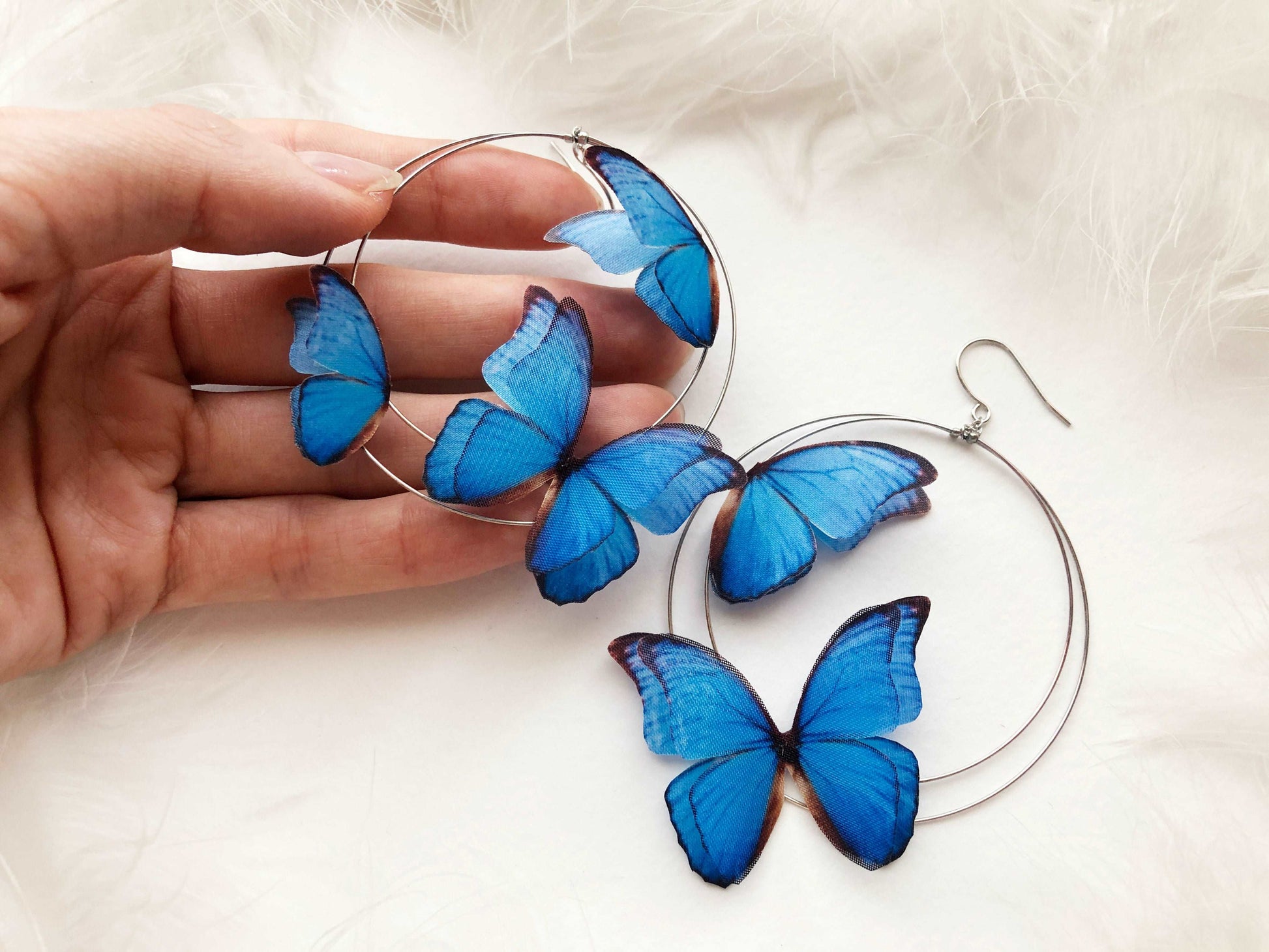 Hand Holds Silver Hoop Earrings with Blue Butterflies on White Feathers Background