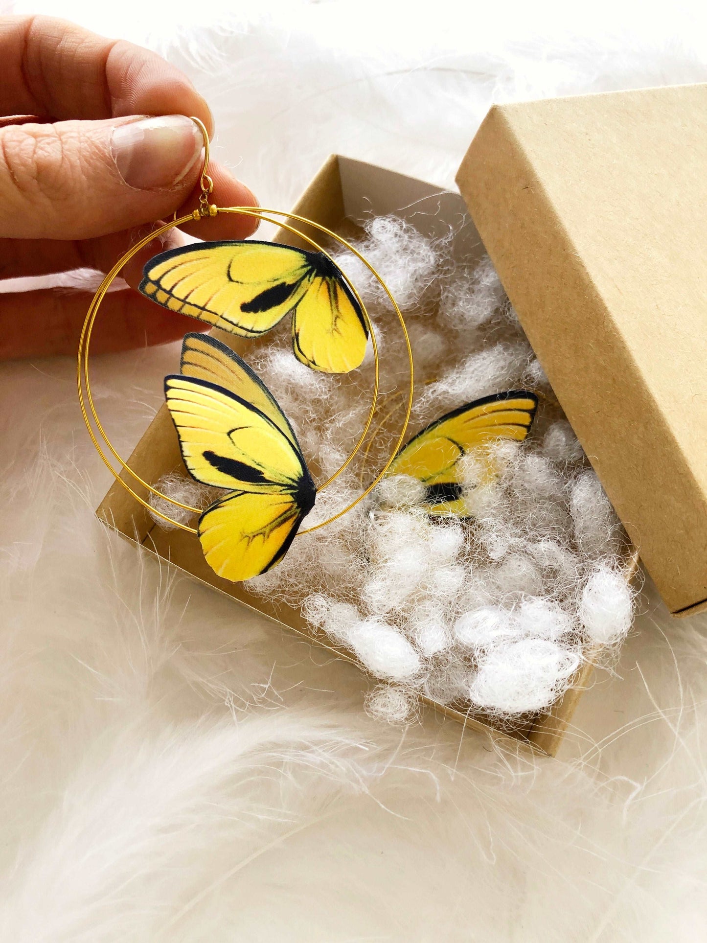 Hand Holds Double Hoop Earrings with Yellow Butterflies in Craft Box on White Feathers