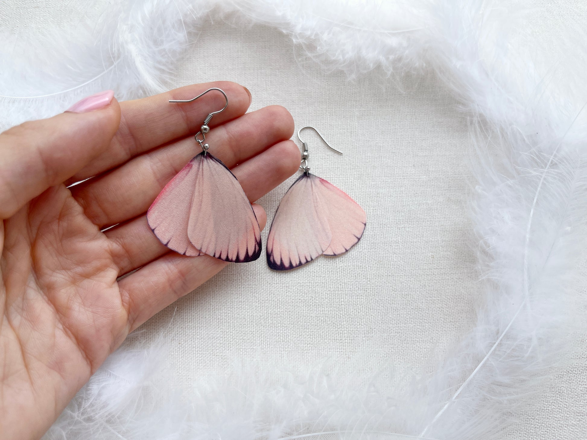 Hand Holds Pink Butterfly Earrings on White Feathers Background